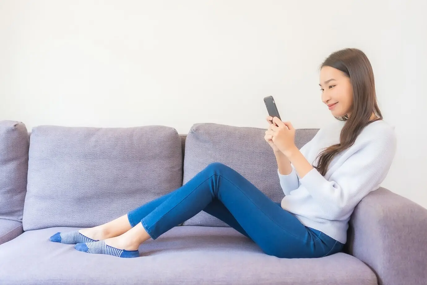 Porträt einer schönen jungen asiatischen Frau, die auf einem Sofa im Innenraum eines Wohnzimmers ein Smartphone benutzt.